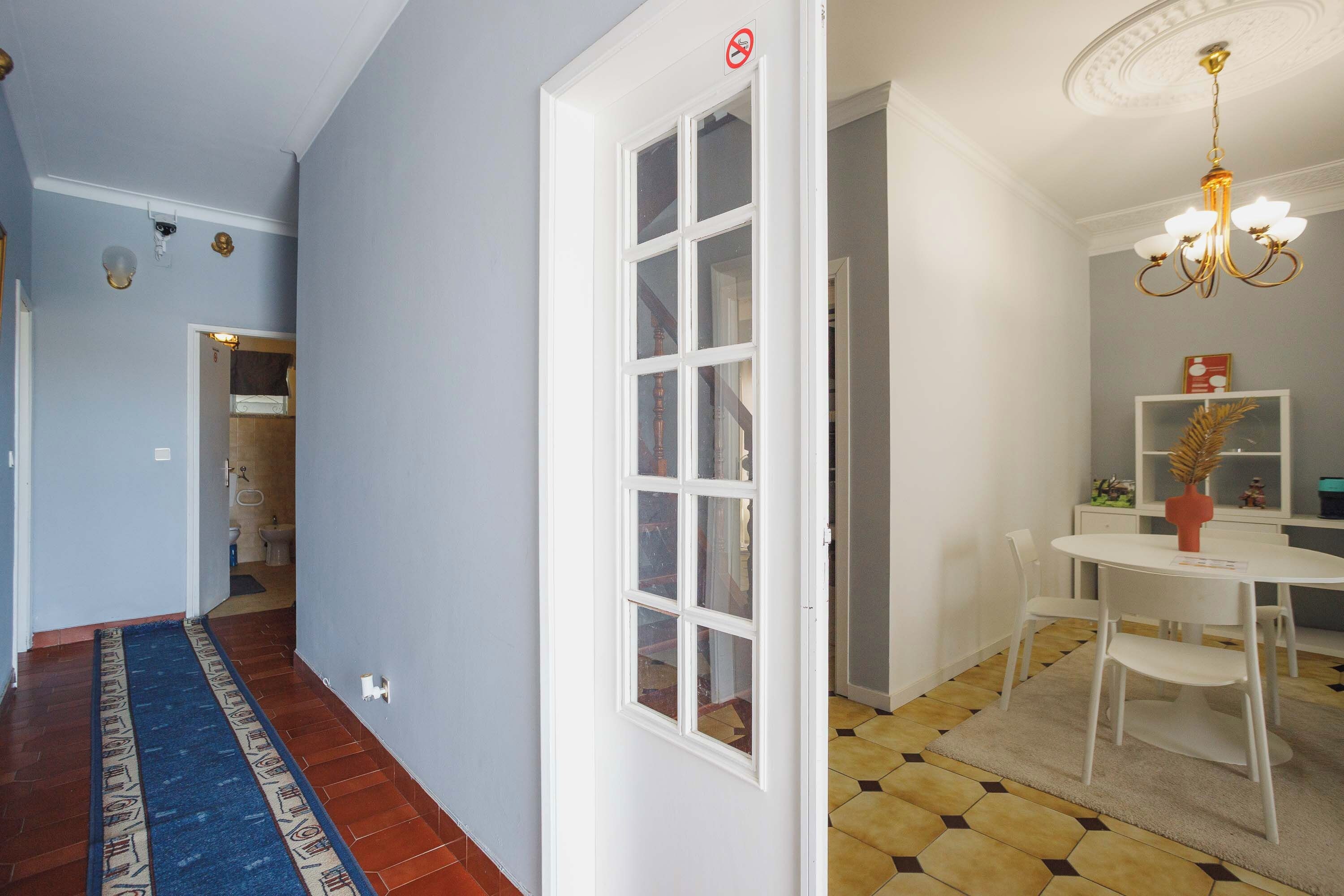 A hallway with a patterned rug, a white door with glass panels, a dining area with a chandelier, and a tiled floor.