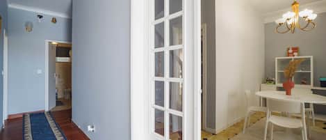 A hallway with a patterned rug, a white door with glass panels, a dining area with a chandelier, and a tiled floor.