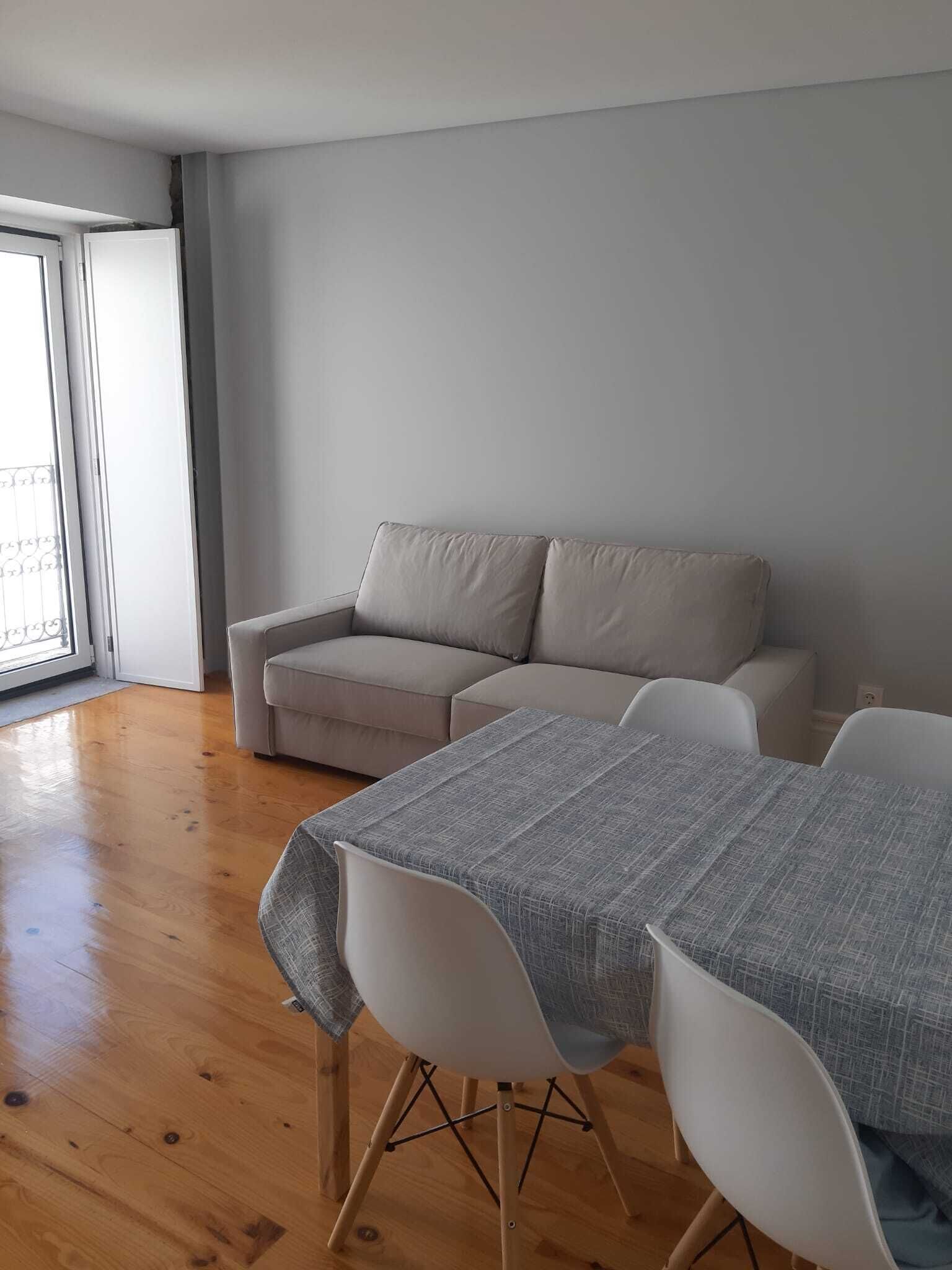 A modern living room with a beige sofa, a dining table with two chairs, and a large window providing natural light.