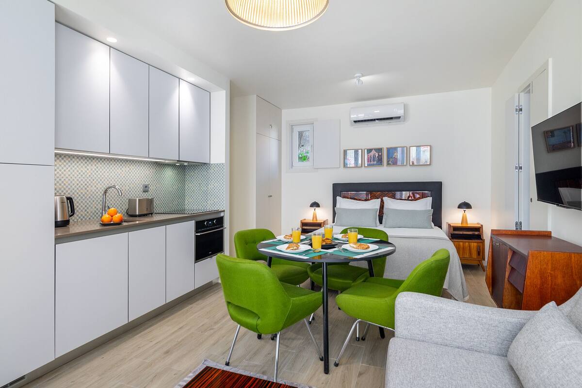 A modern hotel room with a kitchenette, a dining area with green chairs, a bed with a headboard, and a TV mounted on the wall.