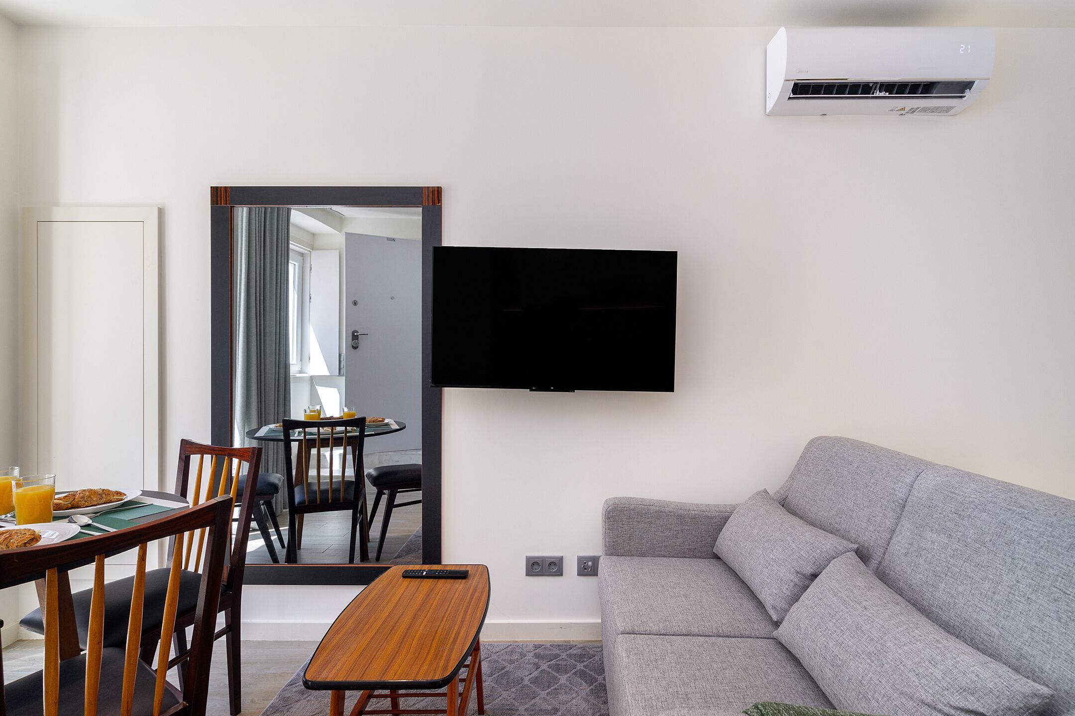 A modern living room with a grey sofa, wooden dining table, and chairs. A flat-screen TV is mounted on the wall, and an air conditioning unit is visible above the sofa.