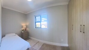 A bedroom with a bed, a nightstand, a lamp, a window, and a wardrobe. - Immaculate 2-bed Apartment in Cape Town (Cape Town)