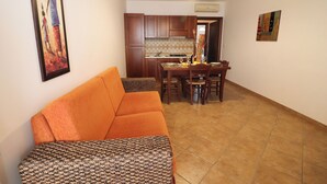 A living room with a sofa, a dining table with chairs, a kitchen area with wooden cabinets, and a hanging artwork. - Samiras Home Cozy Apartment Near the Beach and Otrantos Historic Center (Otranto)