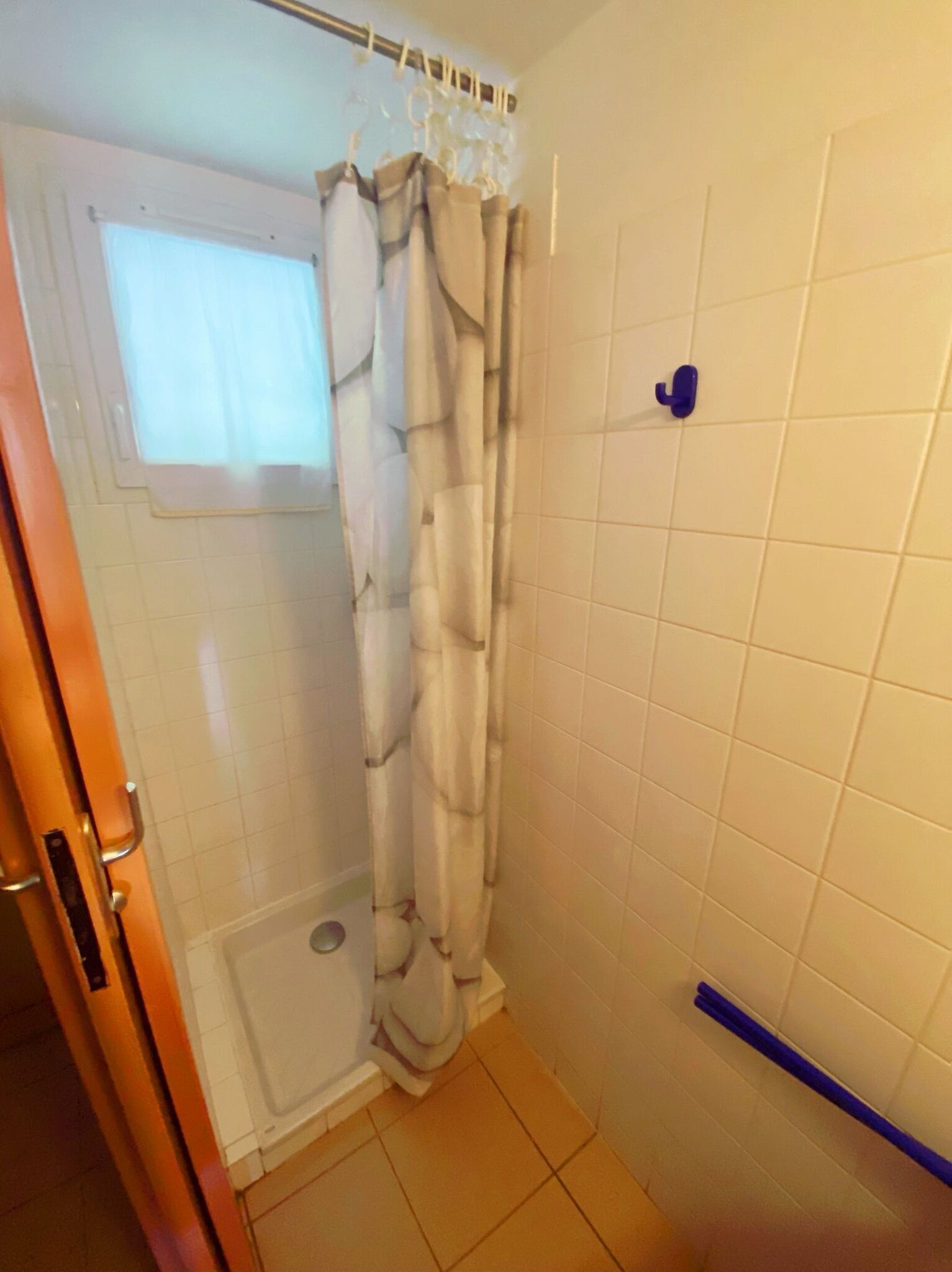 Een kleine, afgesloten douchecabine met een witte betegelde wand, een douchegordijn en een blauwe handgreep.