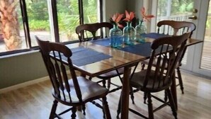 Een eethoek met een houten tafel en stoelen, omgeven door grote ramen met uitzicht op groen.