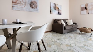 A modern living room with a white dining table, two white chairs, a grey sofa with cushions, and a wicker ottoman. Abstract art is displayed on the walls. - Italianway Comfort - Campania 26 A (Milan)