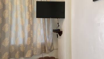 A room with a television mounted on the wall, a wooden bench, a small table with a remote control, and a patterned curtain.