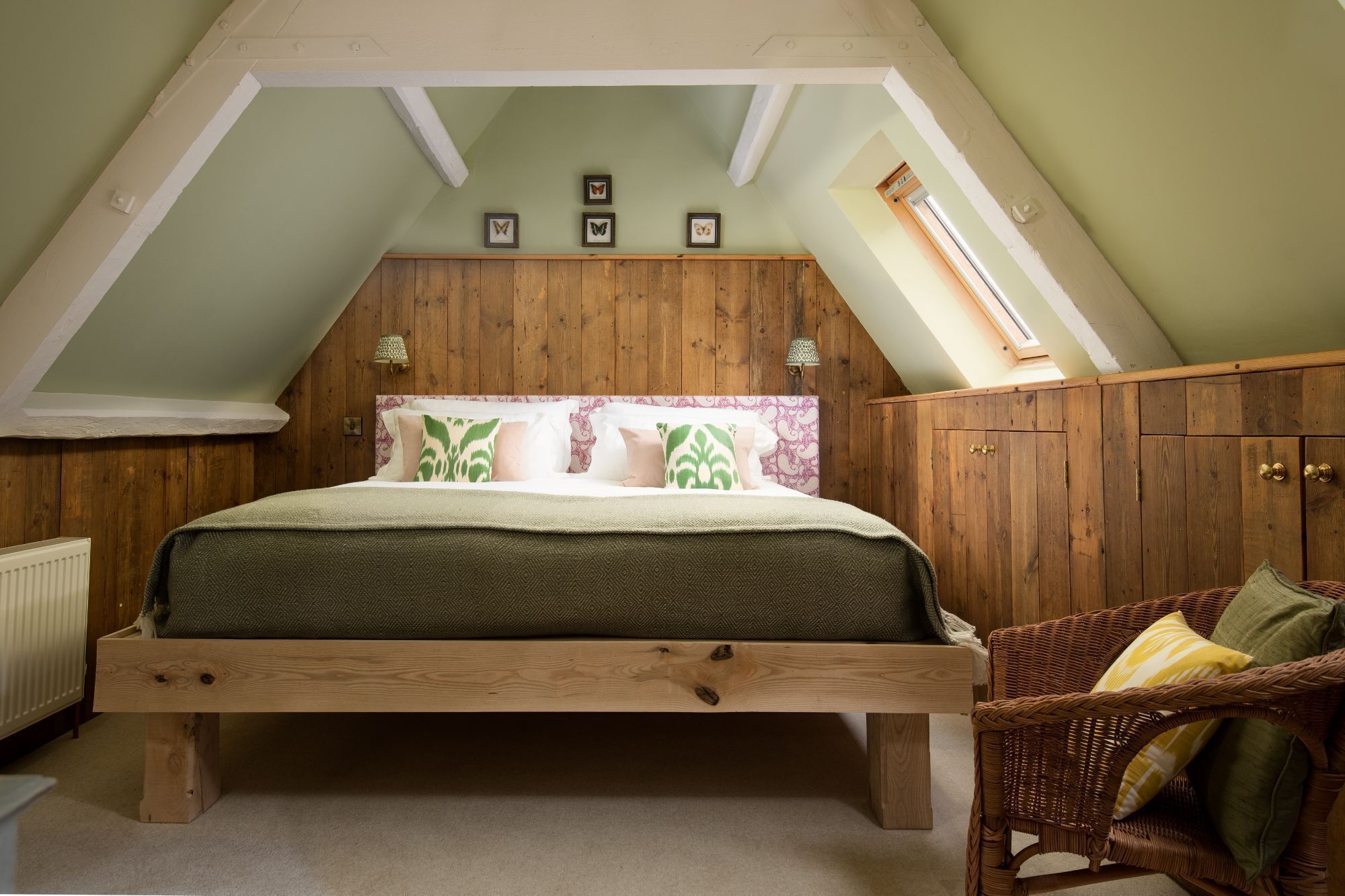 Un dormitorio con techo inclinado, paredes de paneles de madera, una cama con ropa de cama estampada, una silla de mimbre y una ventana pequeña.