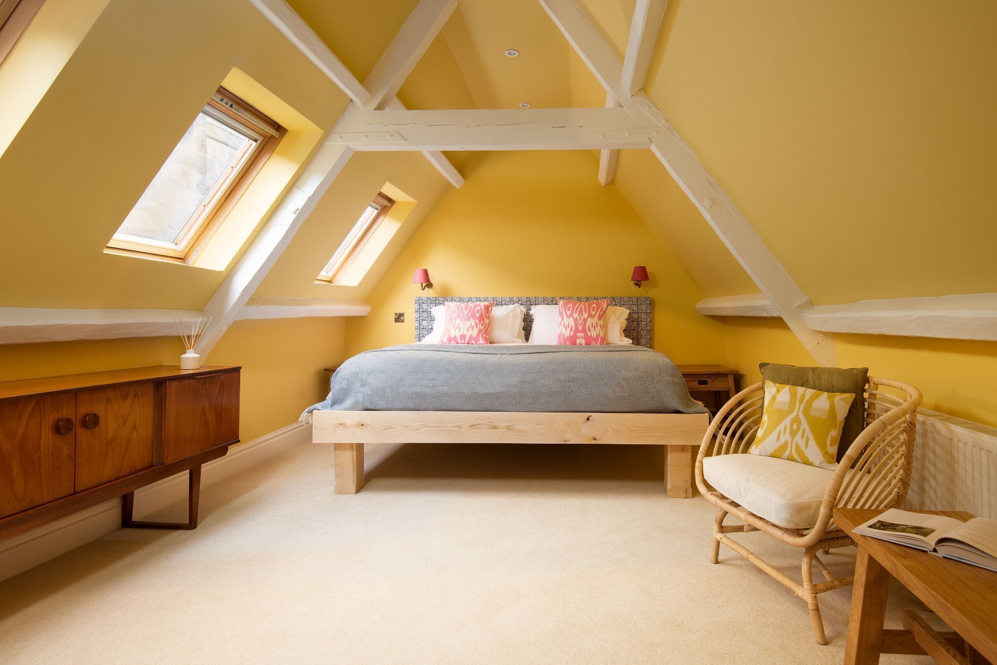 A bedroom with a sloped ceiling, a wooden bed frame, a bedside table, a wicker chair, and a small wooden cabinet.