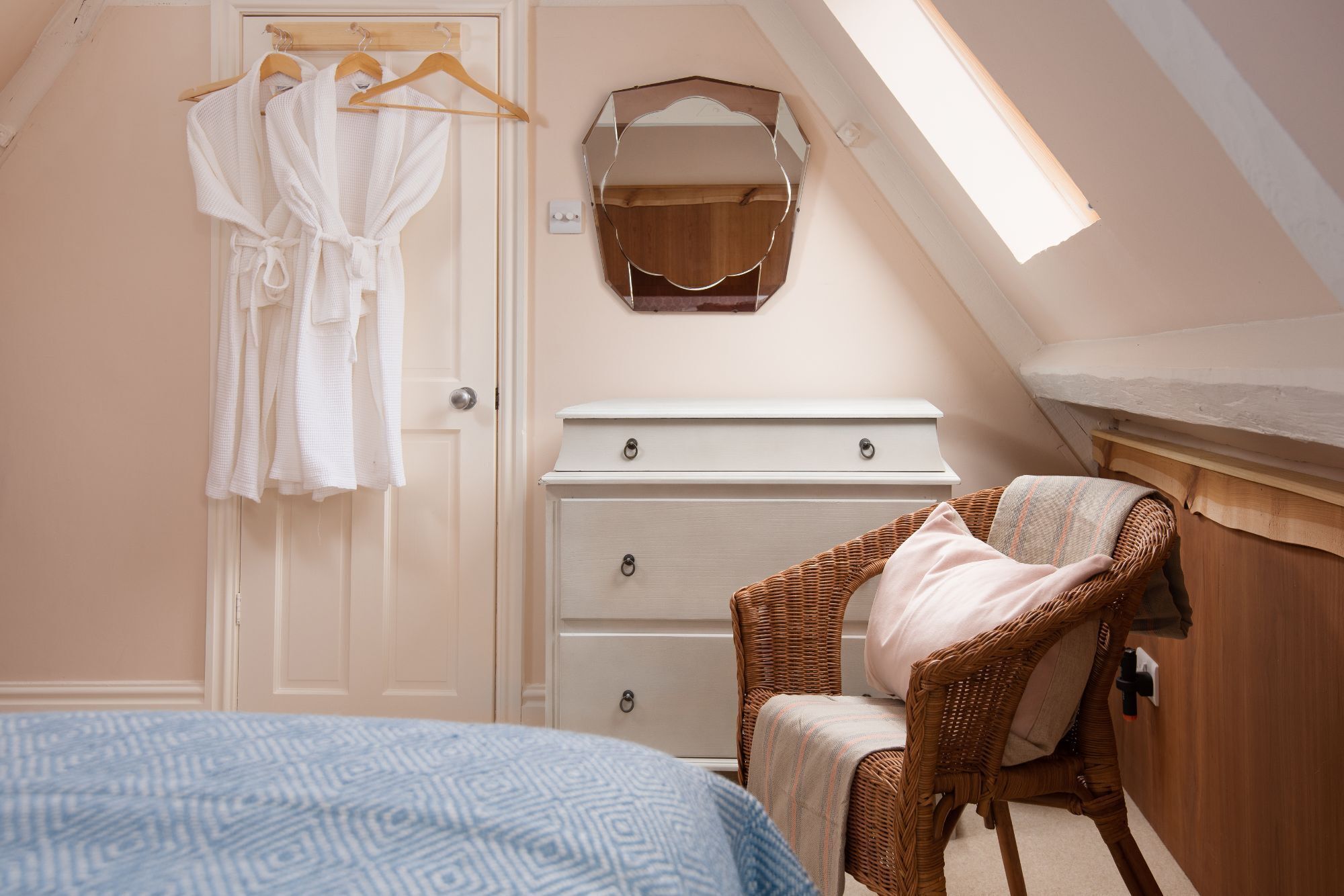 A bedroom with a sloped ceiling, a bed with a patterned blue bedspread, a wicker chair with a pink blanket, a white dresser with drawers, and a wooden mirror on the wall.