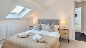 Un dormitorio con tragaluz, una cama con ropa de cama blanca, dos mesitas de noche con lámparas, y vistas a un televisor en la habitación contigua.