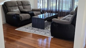 A modern living room with a dark wood floor, a black leather sofa, a black leather armchair, and a glass coffee table. - Pakenham Villa (Pakenham)