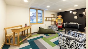 Een kinderspeelruimte met een raam, een houten tafel, een relax fauteuil, een tafelvoetbaltafel en een grote decoratie van een tekenfilmfiguur.