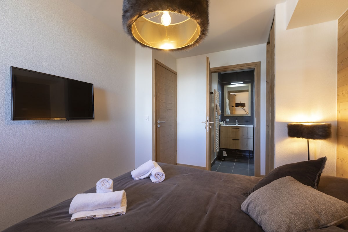 A hotel room with a bed, a flat-screen TV mounted on the wall, a door leading to a bathroom, and a lamp on the bedside table.