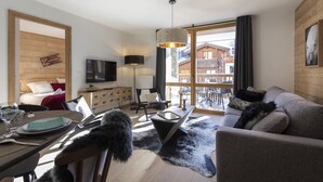 Een moderne woonkamer met een bank, een eettafel en een televisie. Er is een houten wand en een raam met uitzicht op een gebouw.