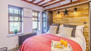 Un dormitorio con cabecera de madera, cama con muchos cojines, ventana con persianas y una bandeja con cruasanes y jugo.