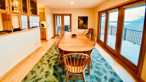A spacious dining area with a long wooden table and chairs, a kitchen with wooden cabinets, and large windows offering a view of a balcony. - Fall Color Specials, near Apple Orchards, with Lake & Island Views (Bayfield)