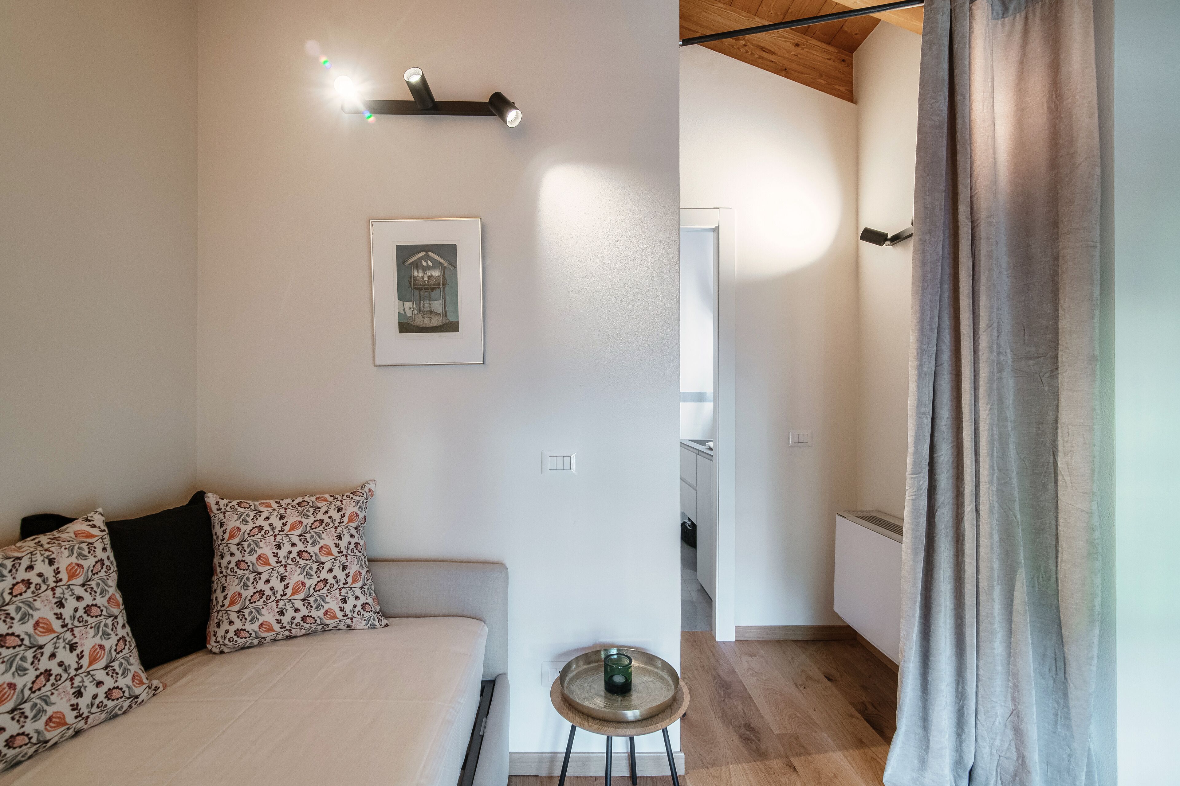 A bedroom with a bed, a small table, a framed picture on the wall, and a view into the kitchen.