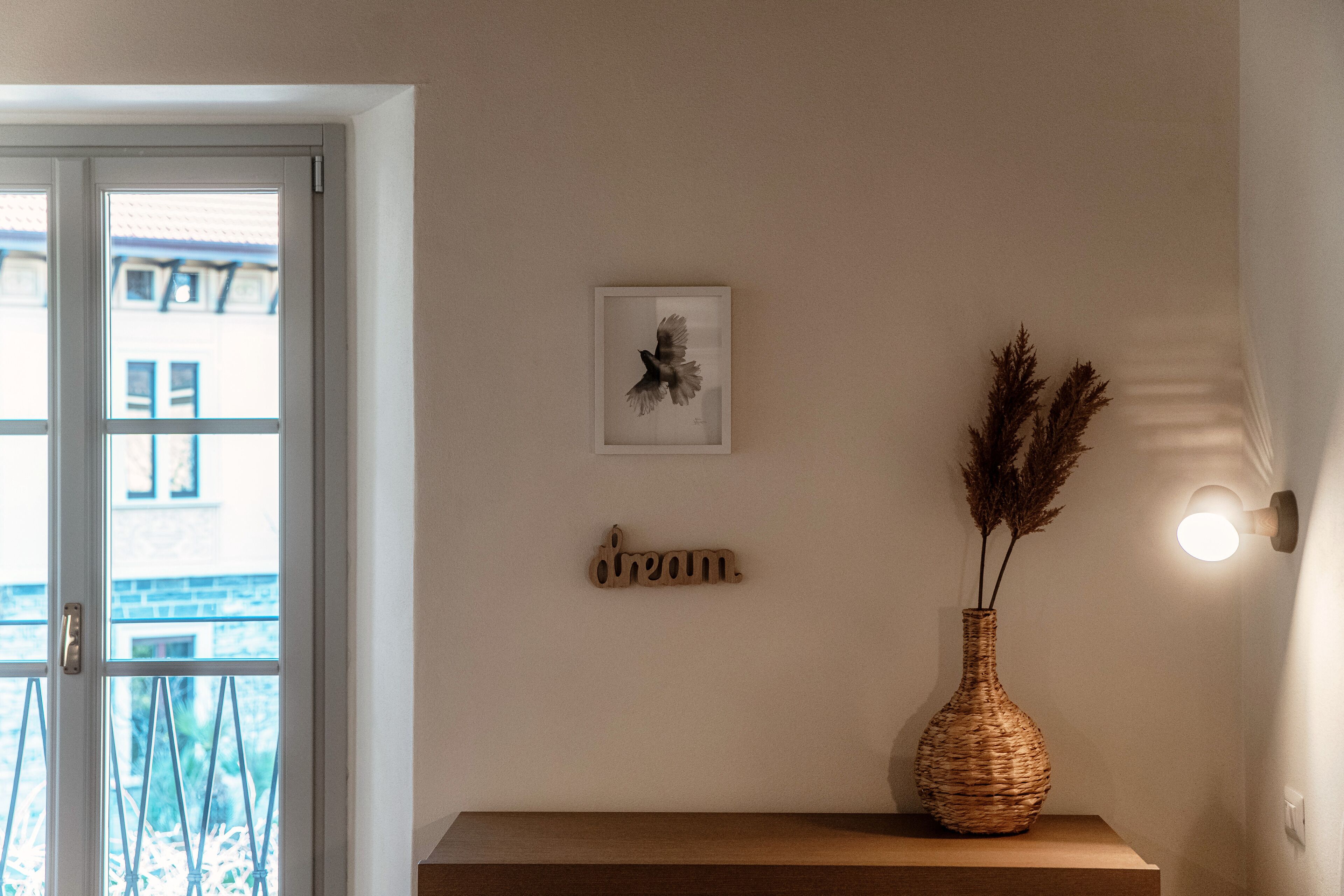 A room with a wooden bench, a vase with dried plants, a framed picture, and a wall-mounted light fixture.
