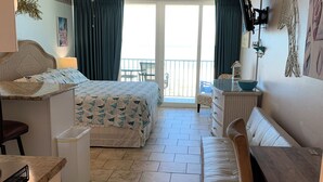 A bedroom with a bed, a desk, a chair, a dresser, a ceiling fan, and a balcony with a view. - Ocean Front Studio Condo on Treasure Island (Treasure Island)
