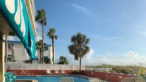 A pool area with lounge chairs, palm trees, and a building with a teal awning. - Ocean Front Studio Condo on Treasure Island (Treasure Island)