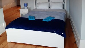 A bedroom with a white bed, a blue blanket, and a nightstand with a lamp.
