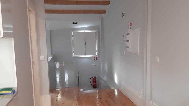 A hallway with wooden flooring, a white wall, and a white ceiling with wooden beams. There is a white door at the end of the hallway, a fire extinguisher mounted on the wall, and a white couch in the foreground.