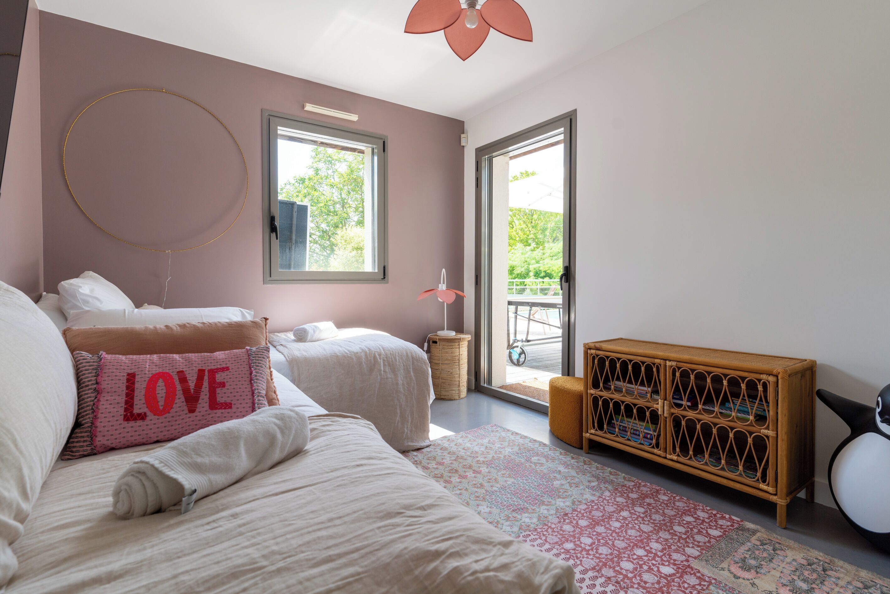 A bedroom with two beds, a large window, a wooden cabinet, and a view of greenery outside.