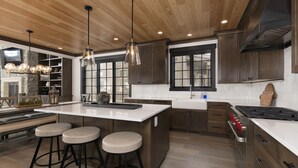 A modern kitchen with a large island, bar stools, wooden cabinets, and pendant lighting.