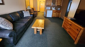 A compact living space with a sofa, a wooden coffee table, a TV stand, a kitchenette with a microwave and refrigerator, and a door leading to another room. - Pine Peaks Lodge & Suites (Crosslake)