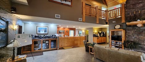 A spacious lobby with a high ceiling, wooden beams, a reception desk, a kitchen area, and a seating area with a television.