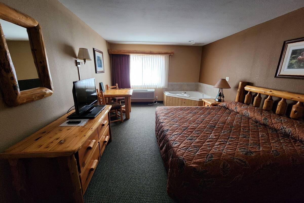 A hotel room with a bed, a desk with a laptop, a chair, a small table, a window with curtains, a bathroom with a bathtub, and a mirror on the wall.