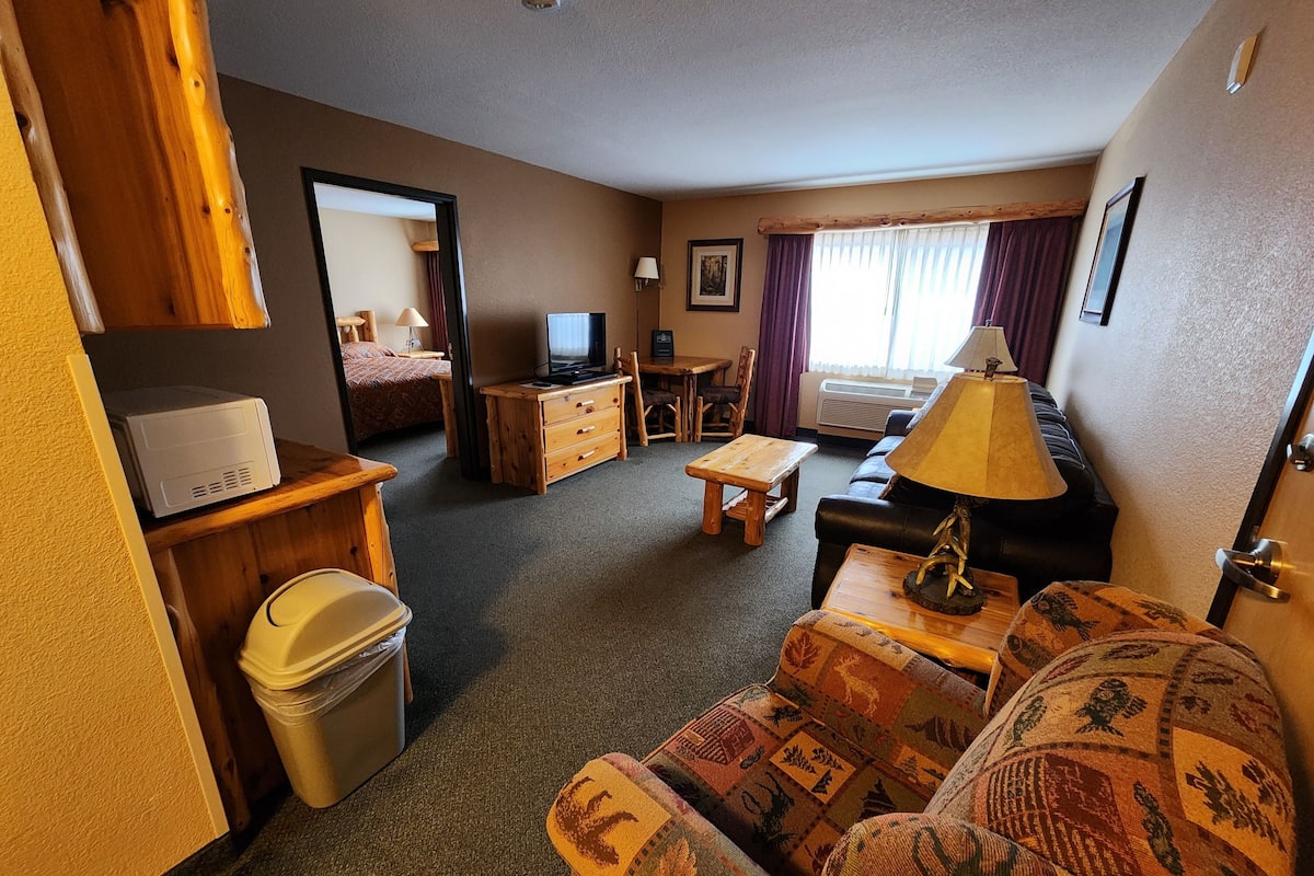 A compact hotel room with a sofa, a small table, a TV, a wooden dresser, and a trash bin.