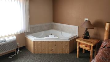 A corner room with a wooden floor, a white bathtub, a small wooden table with a lamp, and a bed with a floral bedspread.