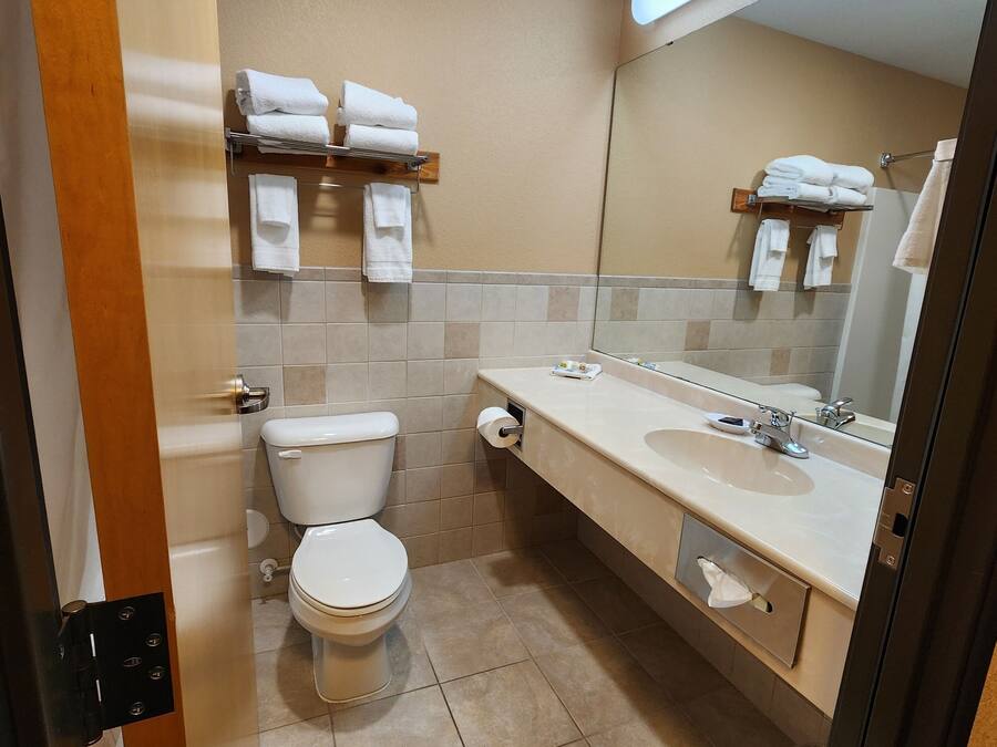 A bathroom with a toilet, sink, and mirror. There are towels on a shelf and a trash can.