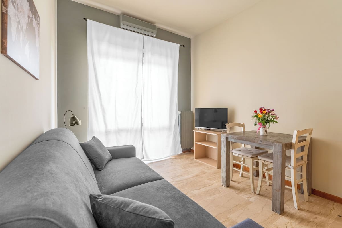 A living room with a grey sofa, a wooden dining table with chairs, a TV, and a window with white curtains.