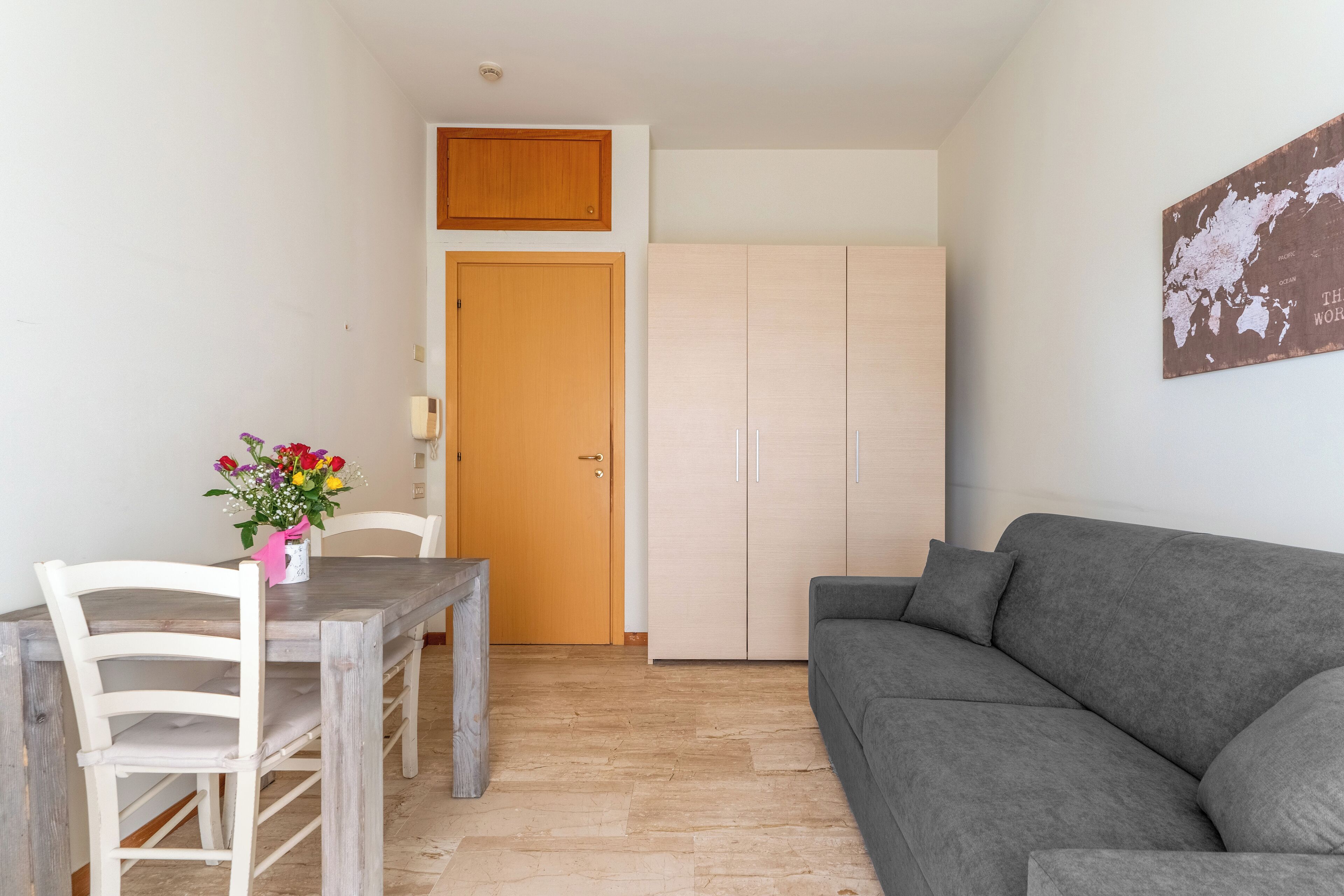 Una sala de estar con un sofá gris, una mesa de comedor de madera con sillas, una puerta de madera y un armario.
