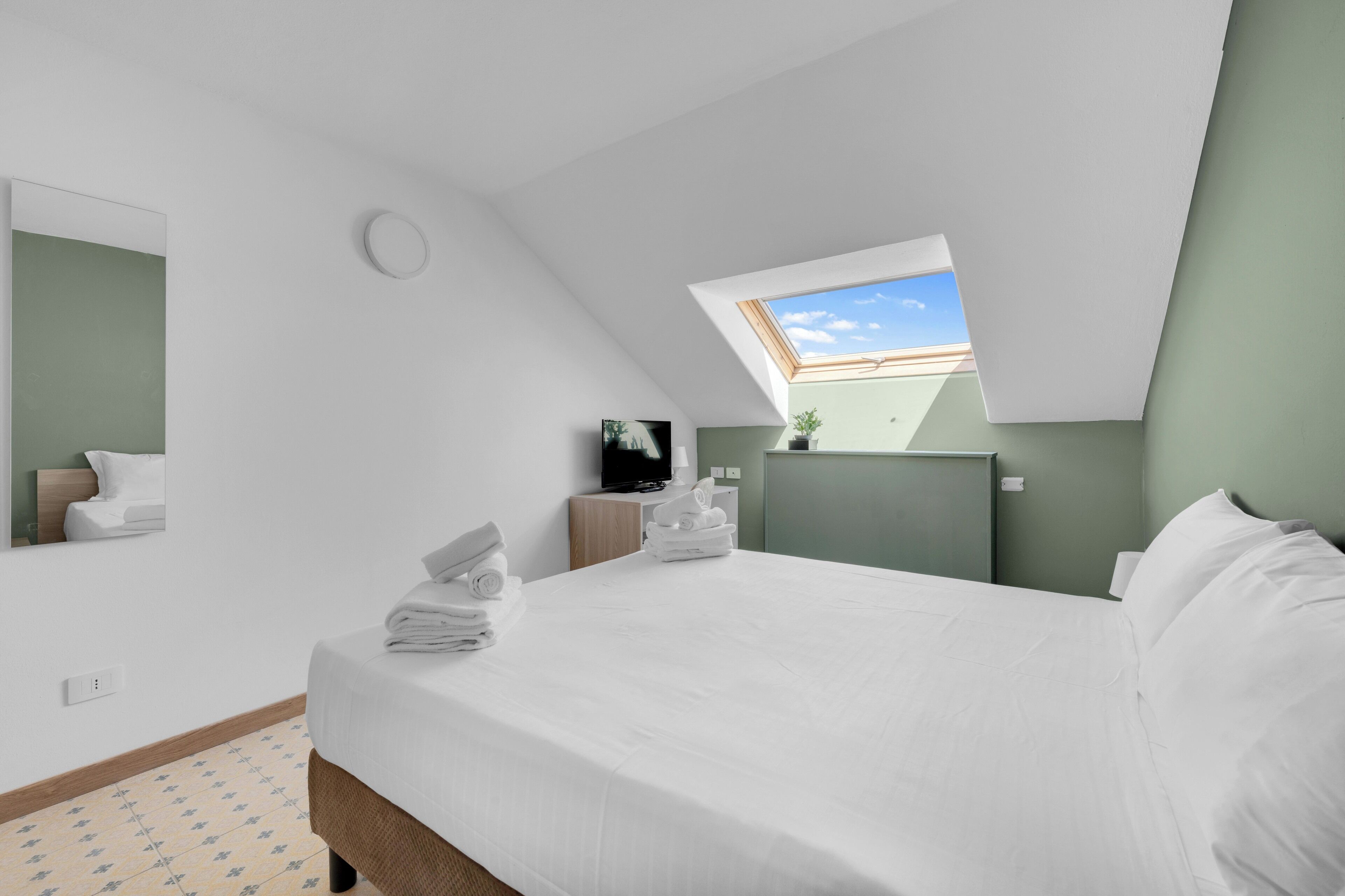 A modern hotel room with a skylight, a bed with neatly folded towels, a small bedside table with a lamp, and a television.