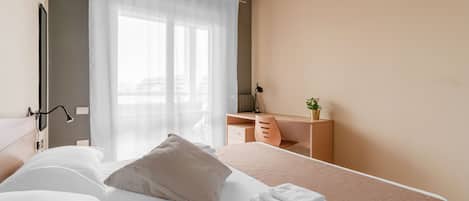A hotel room with a bed, a desk, a chair, and a window with sheer curtains.