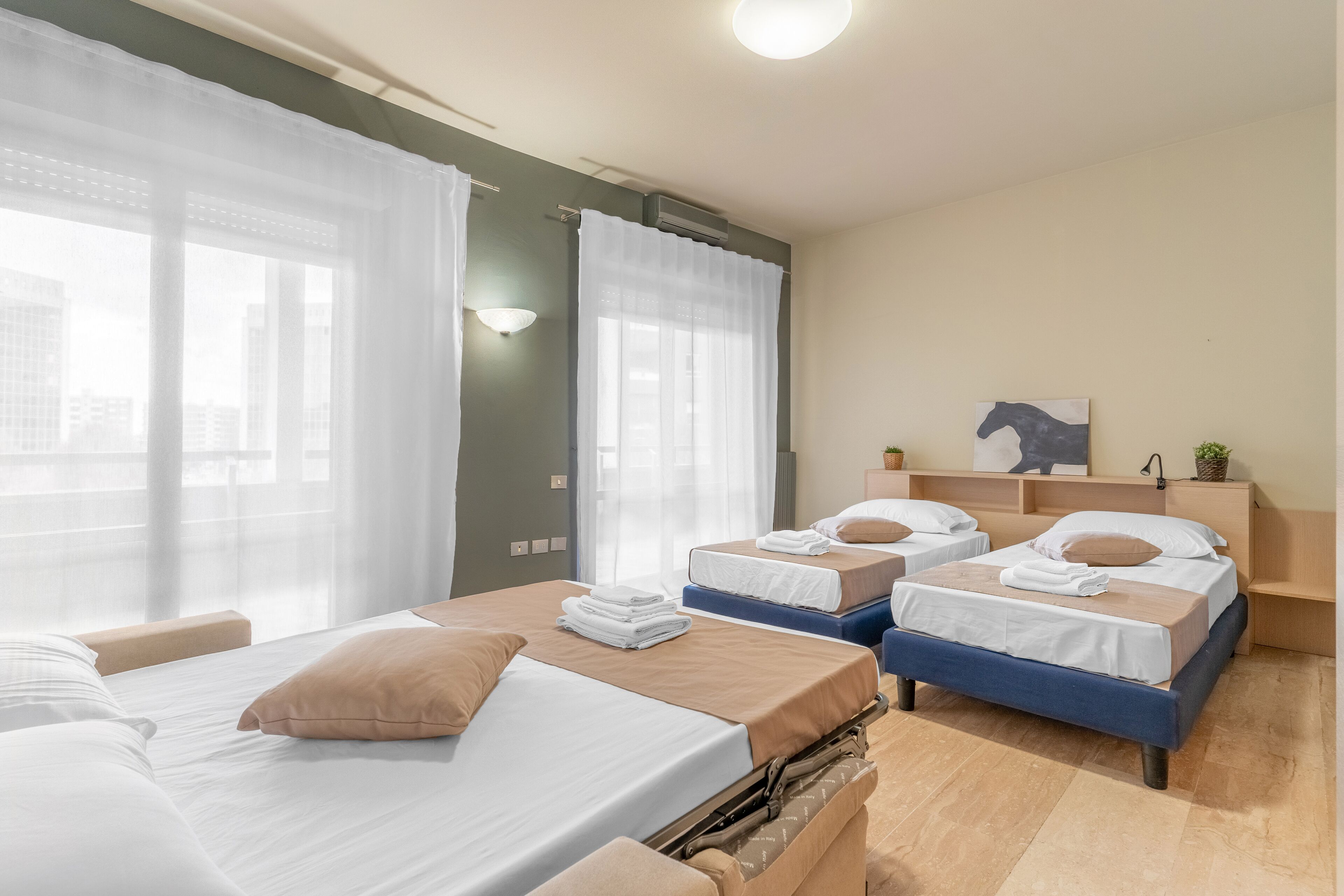 A hotel room with two beds, a large window with sheer curtains, a wall-mounted air conditioner, and a small potted plant.