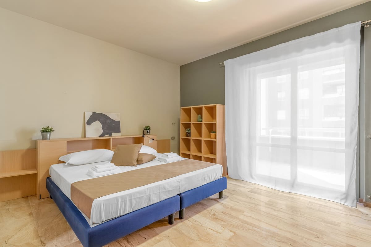 A neatly arranged bedroom with a bed, bedside tables, a bookshelf, and a large window with sheer curtains.