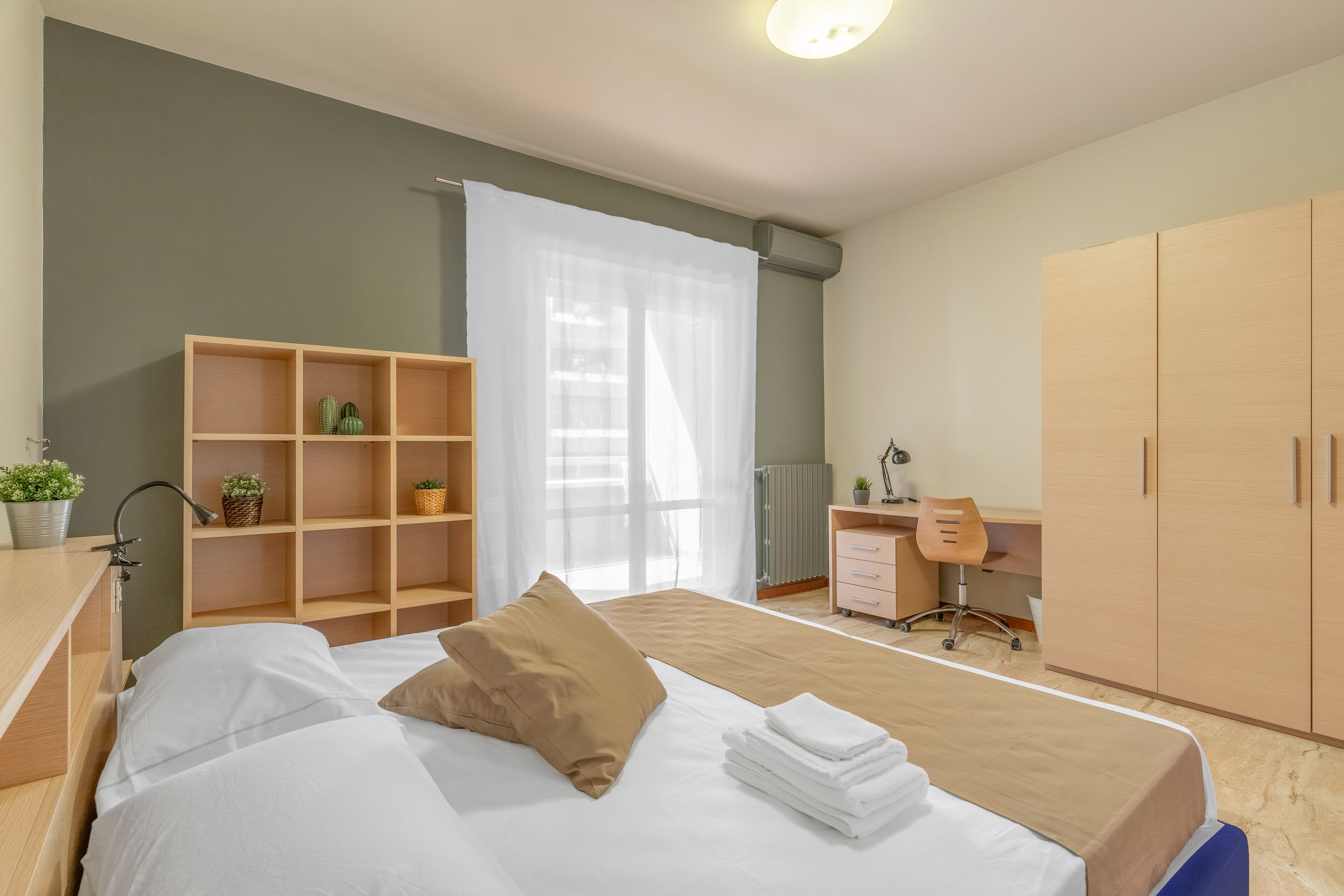 A modern bedroom with a bed, a wooden wardrobe, a desk with a chair, and a small shelf with plants.