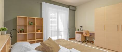 A modern bedroom with a bed, a wooden wardrobe, a desk with a chair, and a small shelf with plants.