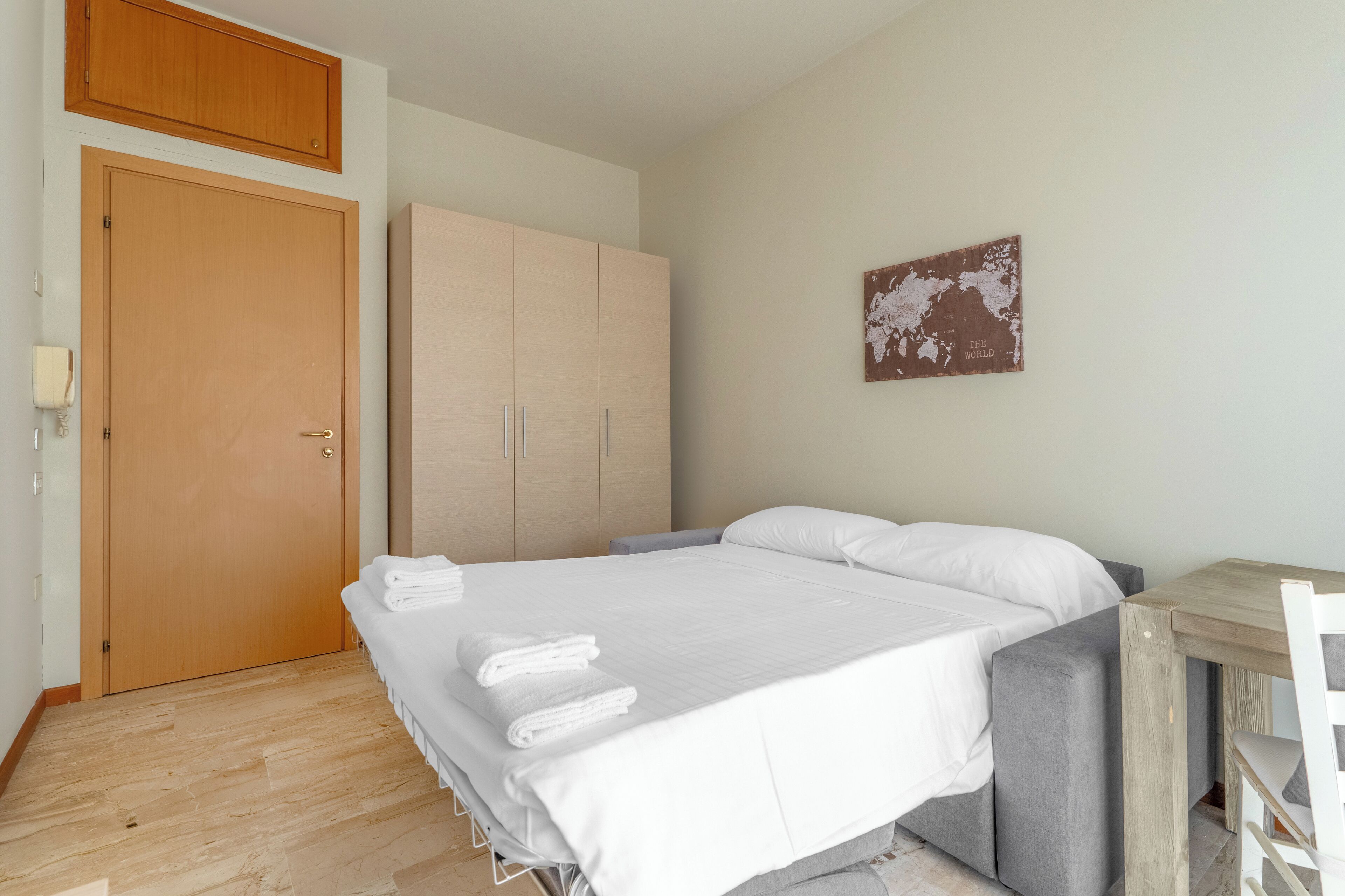 A bedroom with a bed, a wooden wardrobe, a small table, and a chair.