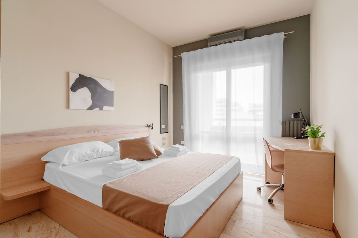 A modern hotel room with a wooden bed, a desk, and a large window with sheer curtains.