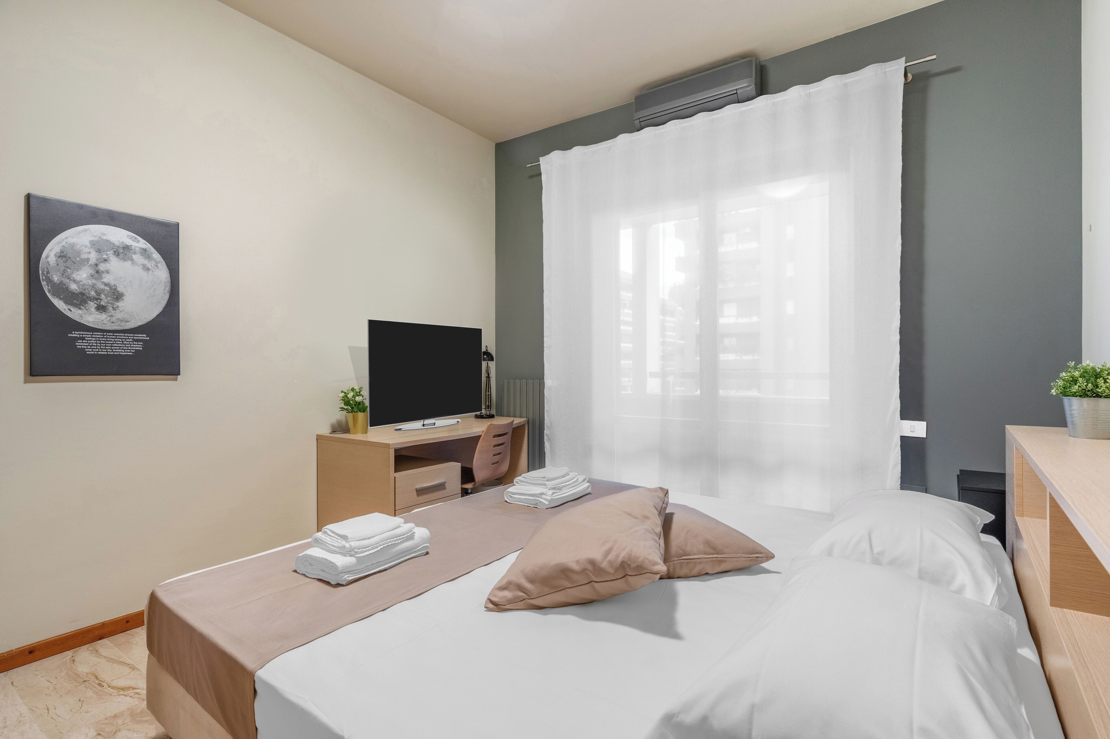 Et hotelværelse med seng, skrivebord med computer, fjernsyn og et vindue med gardiner.