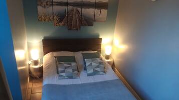 A bedroom with a bed, two bedside tables, a wall-mounted lamp, and a painting above the bed.