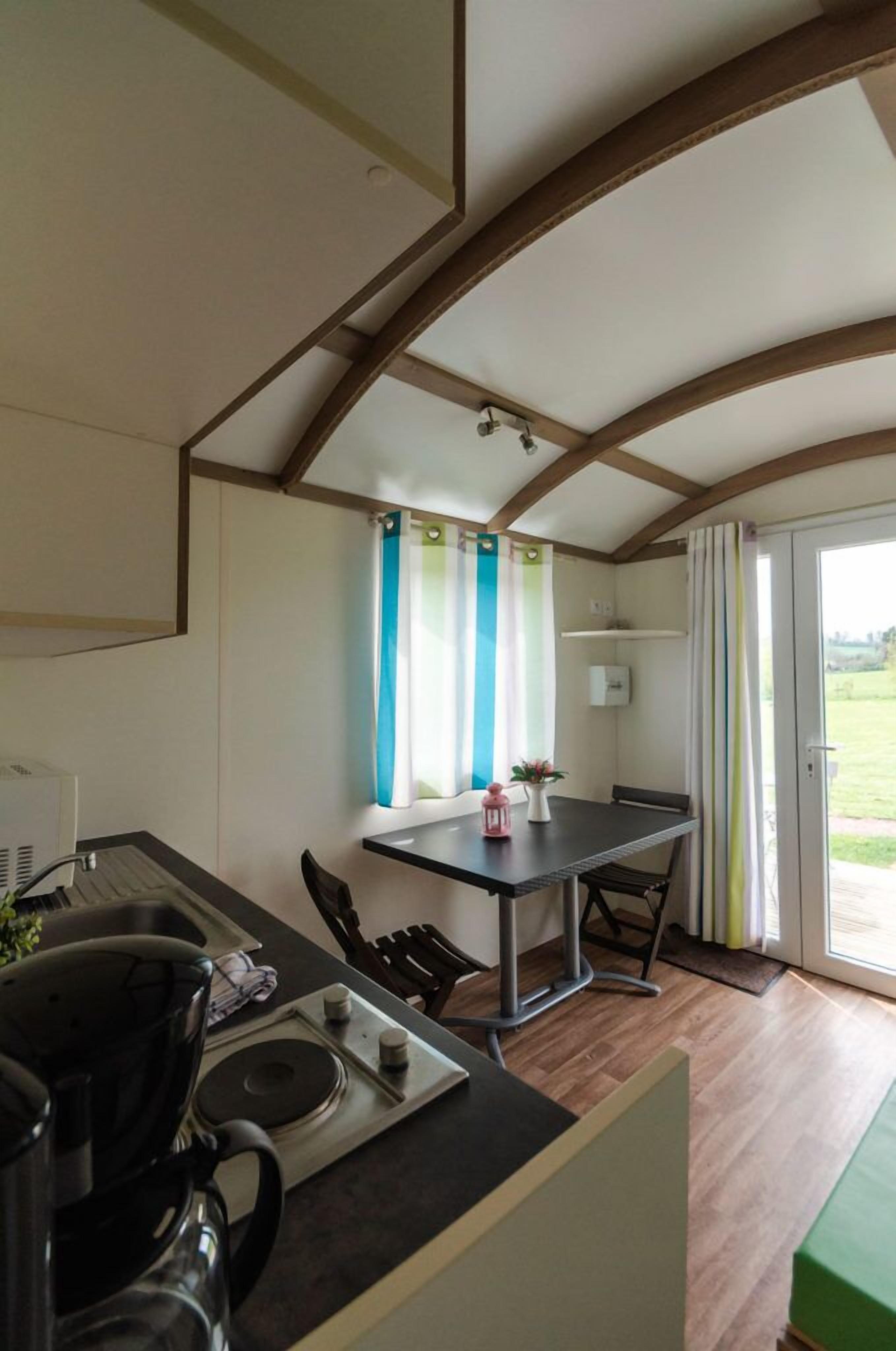A compact kitchen with a dining area, wooden ceiling beams, a window with curtains, and a view of the outdoors.