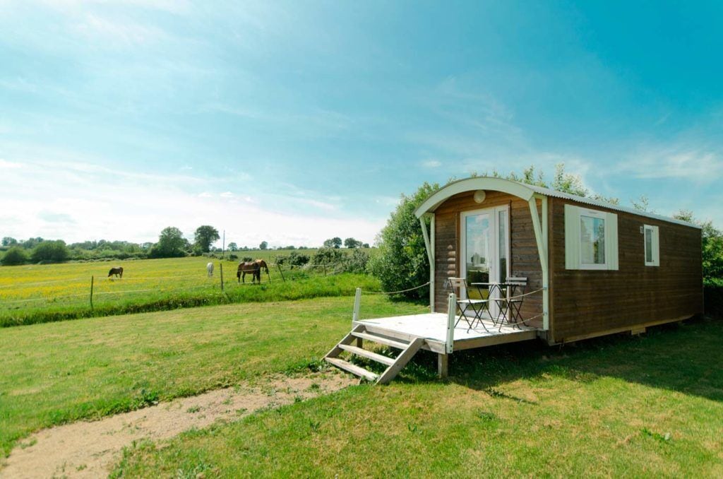 Una cabaña de madera pequeña con un porche y una mesa, rodeada por un campo cubierto de hierba y un caballo a lo lejos.