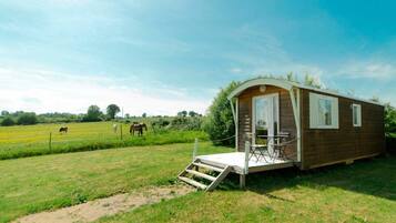 Una cabaña de madera pequeña con un porche y una mesa, rodeada por un campo cubierto de hierba y un caballo a lo lejos.